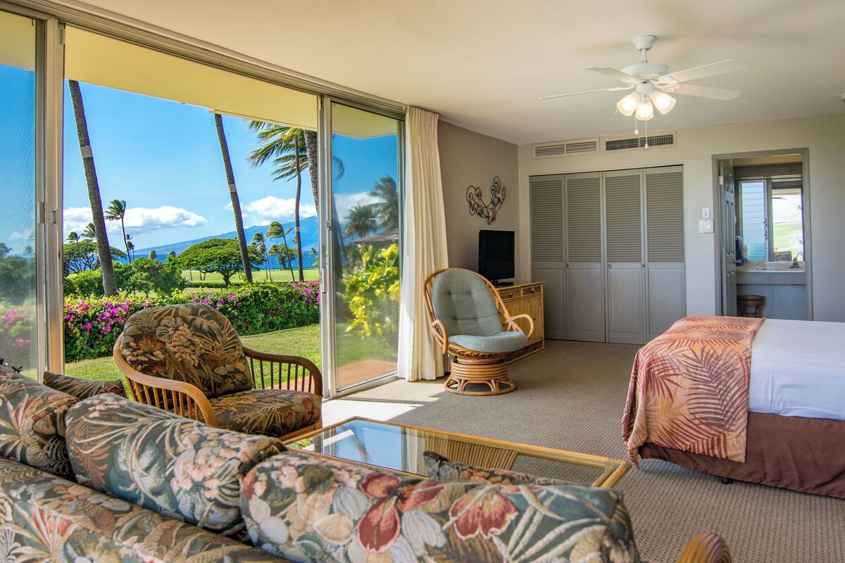 Quarto de hotel com uma grande porta de vidro deslizante que dá para um jardim, uma cama com colcha de tema tropical, uma poltrona de vime e uma televisão sobre um móvel.