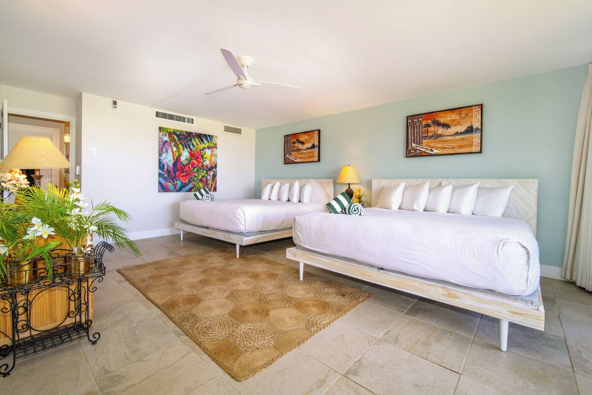 Quarto de hotel com duas camas de solteiro, um ventilador de teto, uma pintura grande e uma planta decorativa.