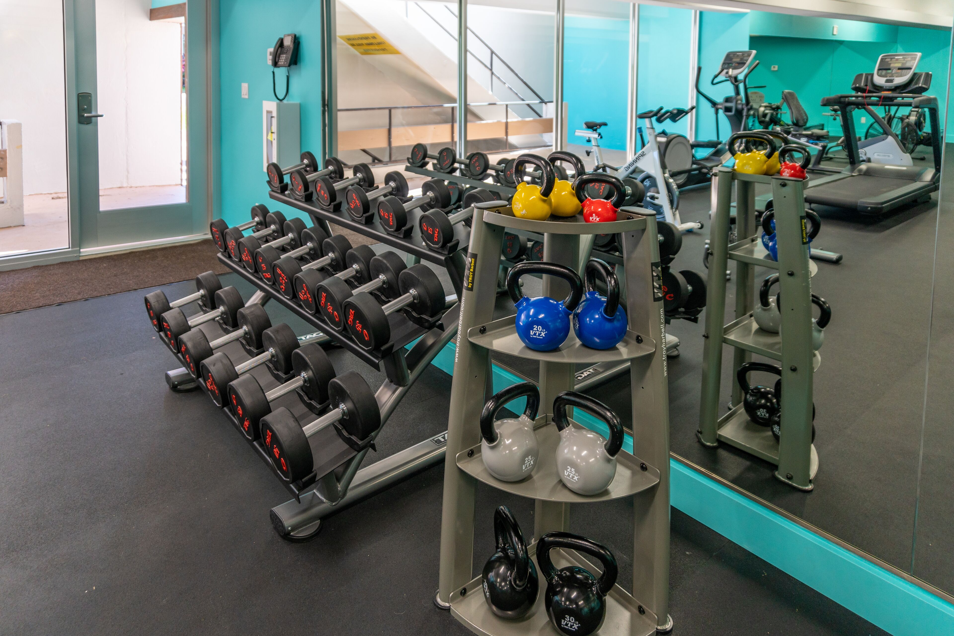 Ein Fitnessstudio mit Gewichthebequipment, darunter Kurzhanteln und Kettlebells, sowie Cardiogeräten.