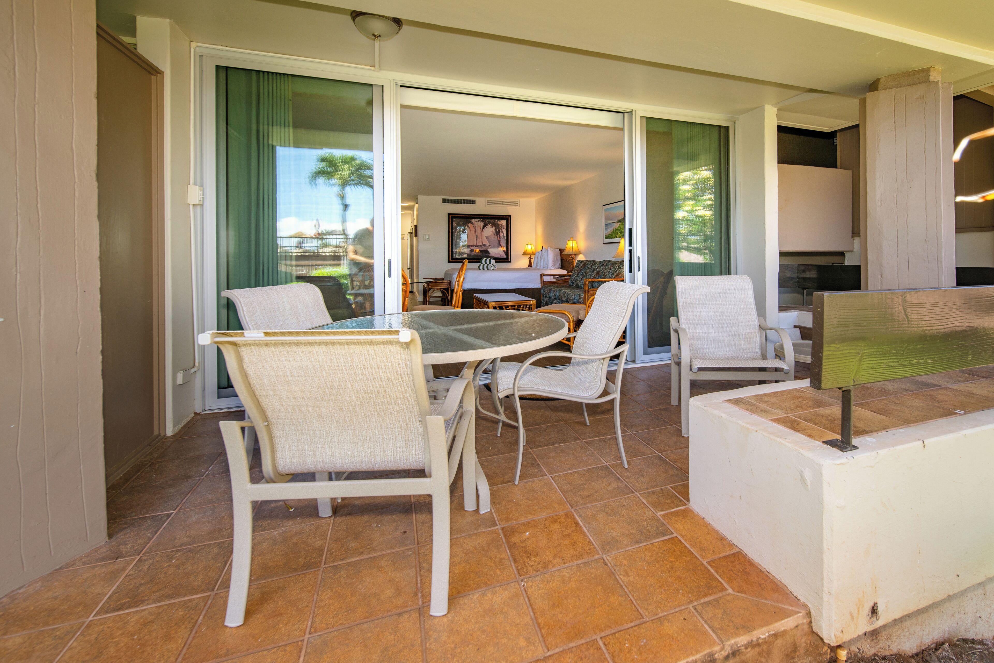 A dining area with a round table and four chairs, a small kitchen area with a stove and cabinets, and a sliding glass door leading to a balcony with a view of palm trees.