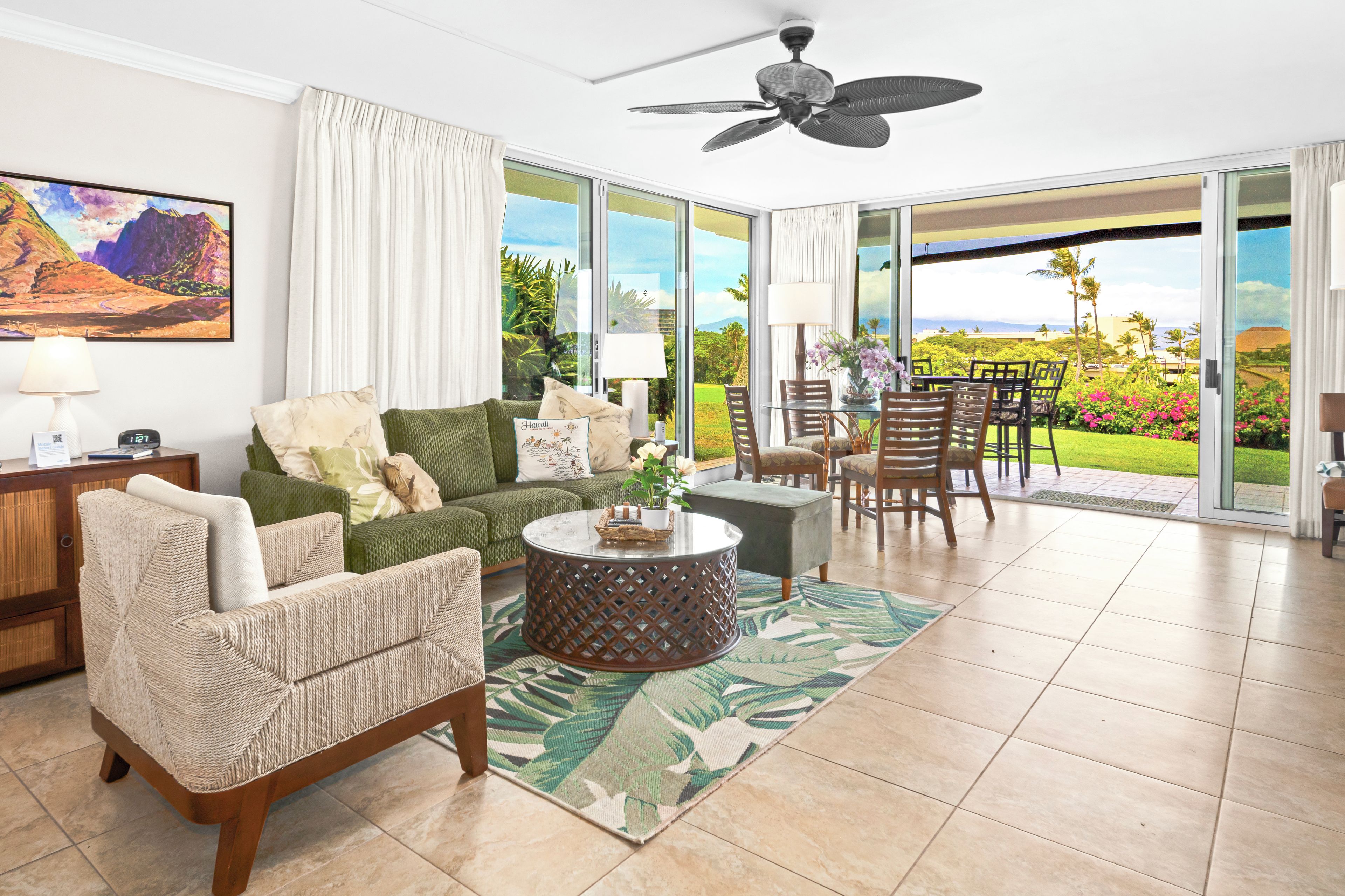 A spacious living area with a green sofa, a wicker chair, a round coffee table, and a view of a lush garden through large sliding glass doors.