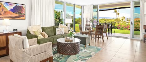 A spacious living area with a green sofa, a wicker chair, a round coffee table, and a view of a lush garden through large sliding glass doors.