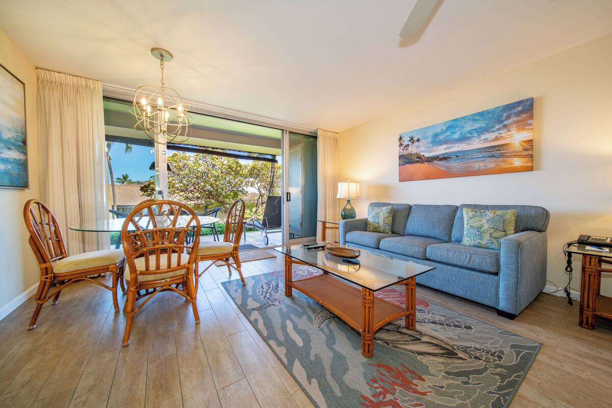 Uma sala de estar com uma mesa de jantar de vidro, cadeiras de vime, um sofá azul e uma pintura de uma cena de praia.