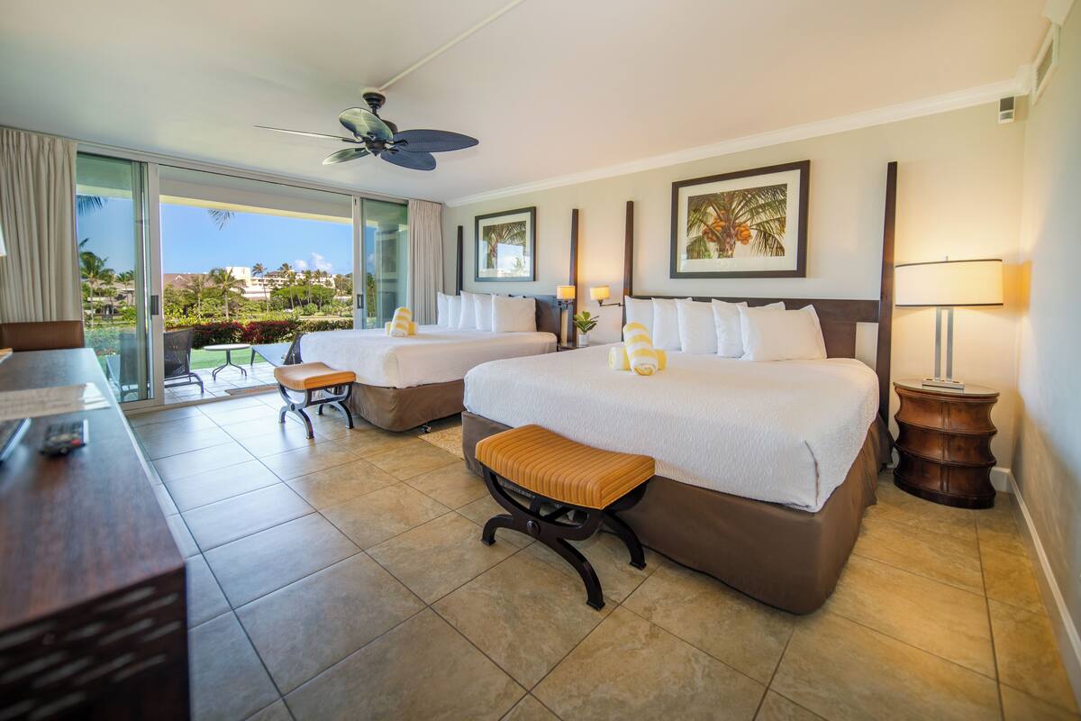 Quarto de hotel com duas camas, uma escrivaninha, uma cadeira, um banquinho, um ventilador de teto e vista para o exterior.