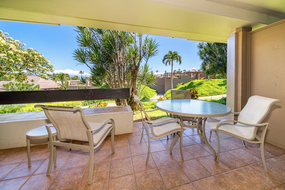 Alpendre coberto com mesa redonda e quatro cadeiras, com vista para um jardim ajardinado com palmeiras e casas vizinhas.