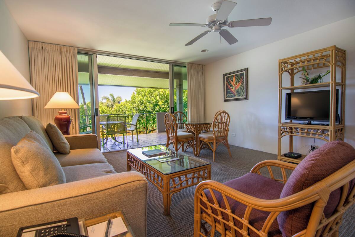 Quarto de hotel com sofá, cadeiras, mesa de centro de vidro, televisão sobre suporte de madeira e varanda com vista para área verde.