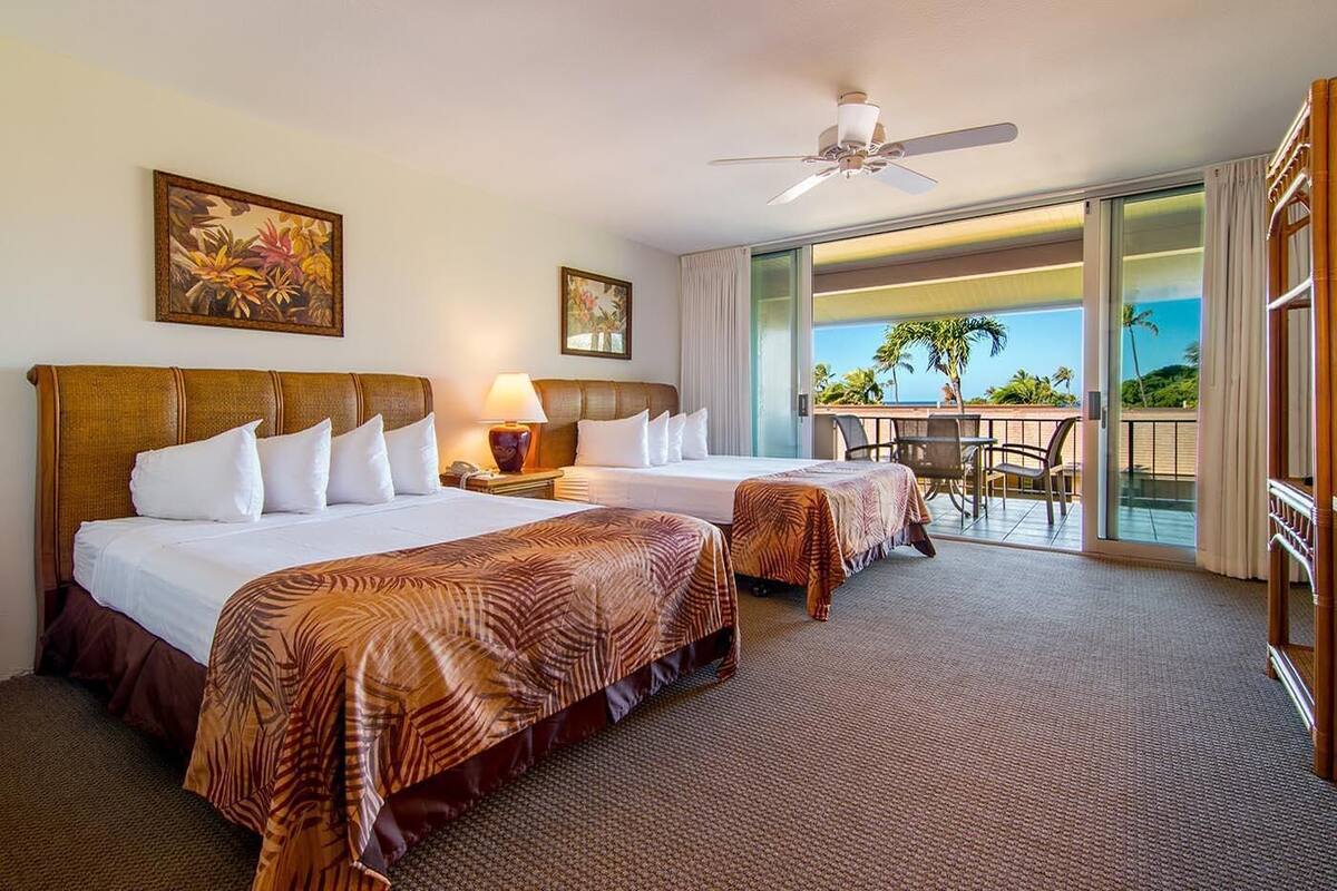 Ein Hotelzimmer mit zwei Betten, einem Deckenventilator, einem Balkon mit Tisch und Stühlen sowie Blick auf den Ozean.