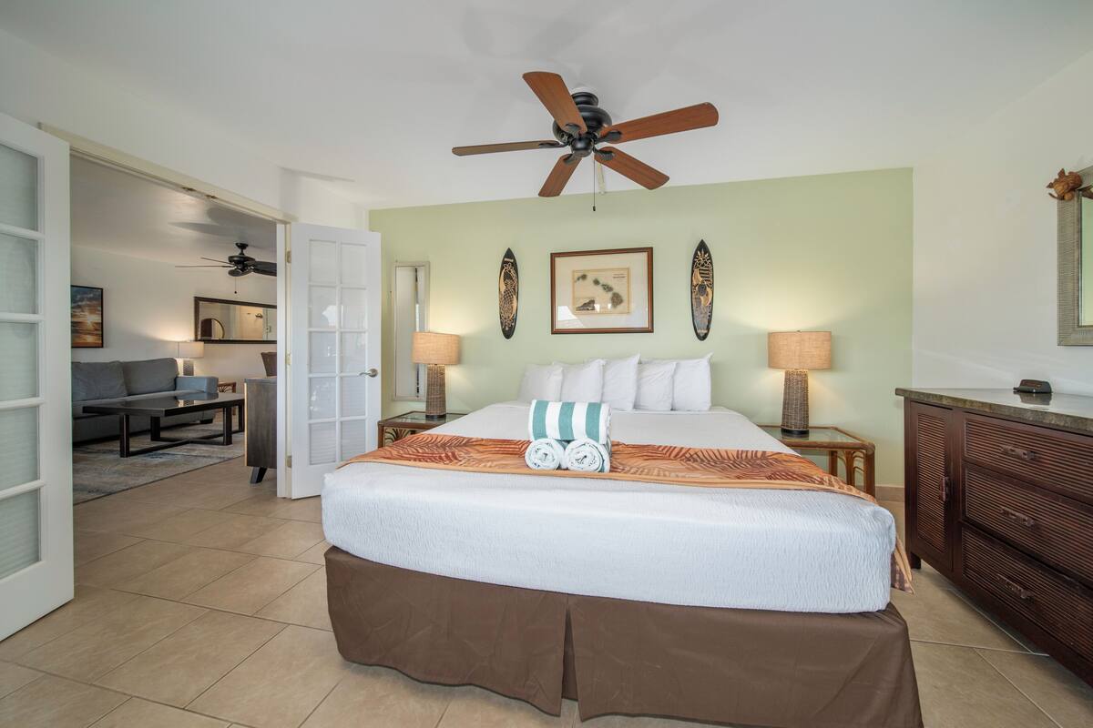 Quarto de hotel com uma cama grande, um ventilador de teto, uma mesa com cadeira e uma área de refeições visível por uma porta aberta.