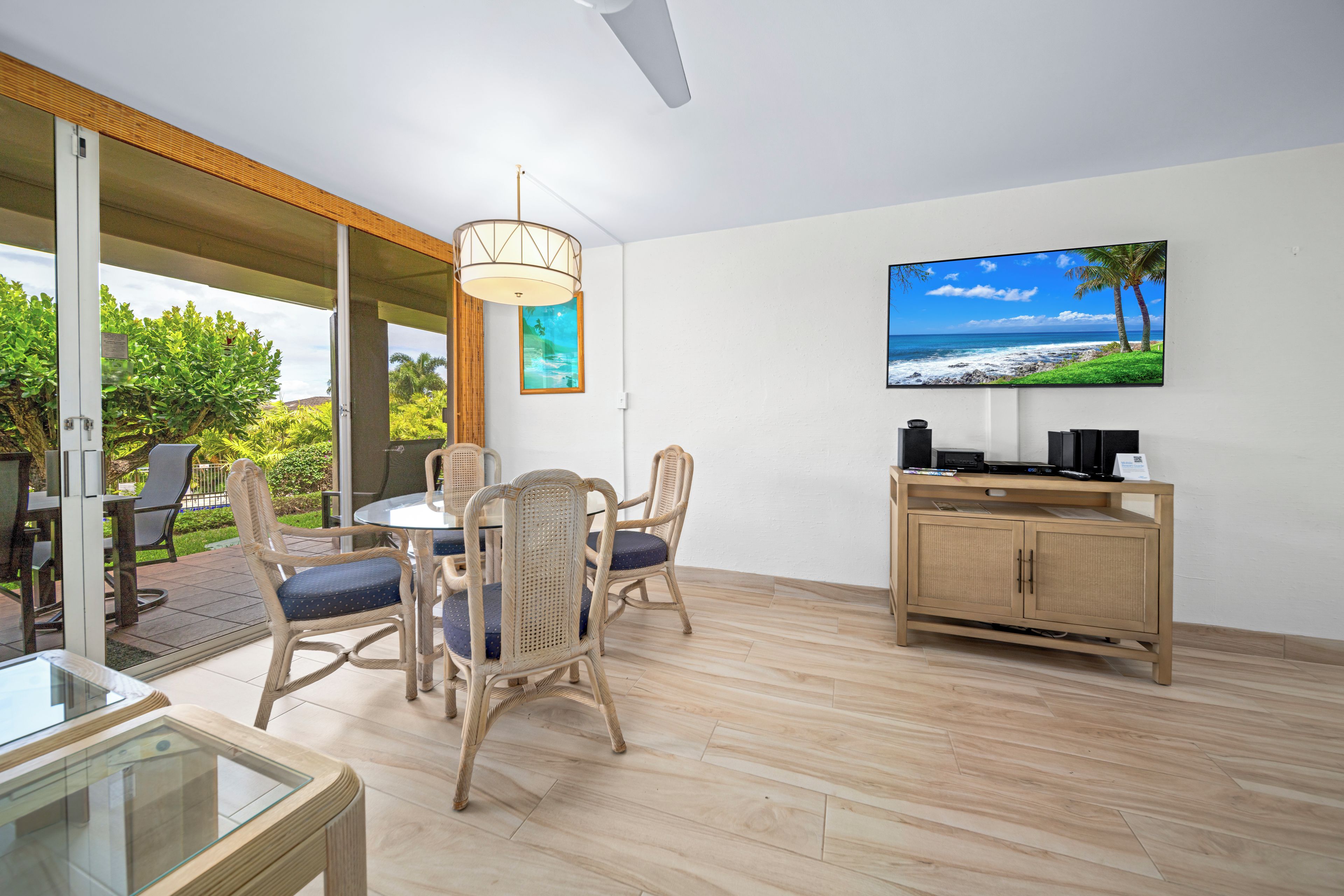 A modern living room with a glass table, wicker chairs, a wooden cabinet, and a large TV screen displaying a beach scene.