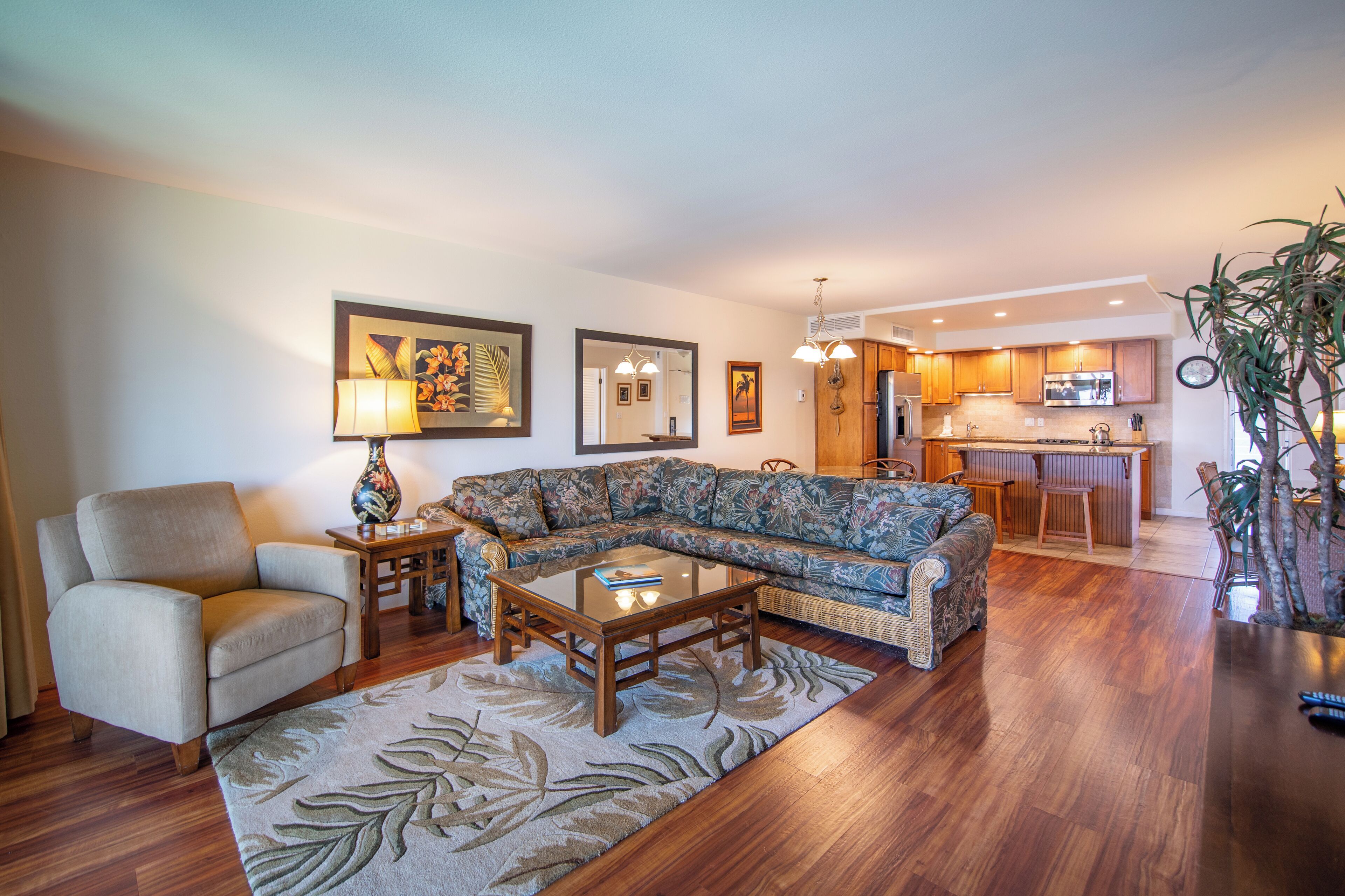 A living room with a sofa, armchair, coffee table, and a rug. There is a kitchen in the background with a microwave and cabinets.
