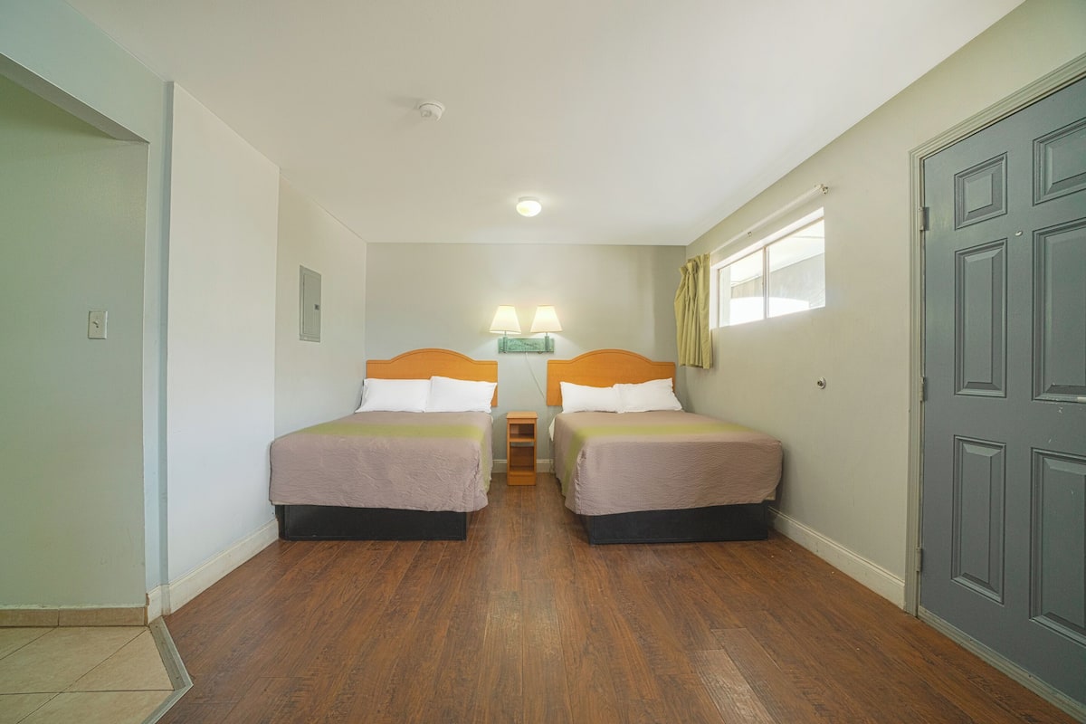 A room with two beds, a nightstand with lamps, a window with curtains, and a door.