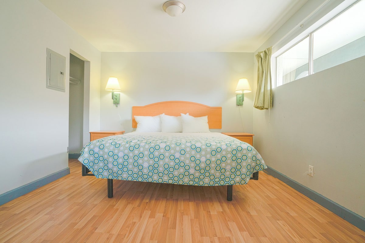 A bedroom with a wooden floor, a bed with a patterned bedspread, two wall-mounted lamps, and a window with curtains.