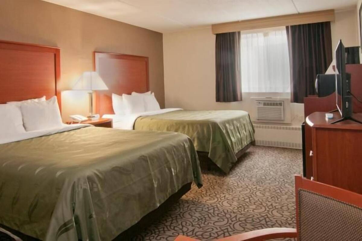 A hotel room with two beds, a desk, a lamp, an air conditioning unit, and a window with curtains.