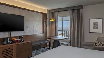 A hotel room with a large bed, a desk with a lamp, a chair, a small table, a TV, and a balcony with curtains.