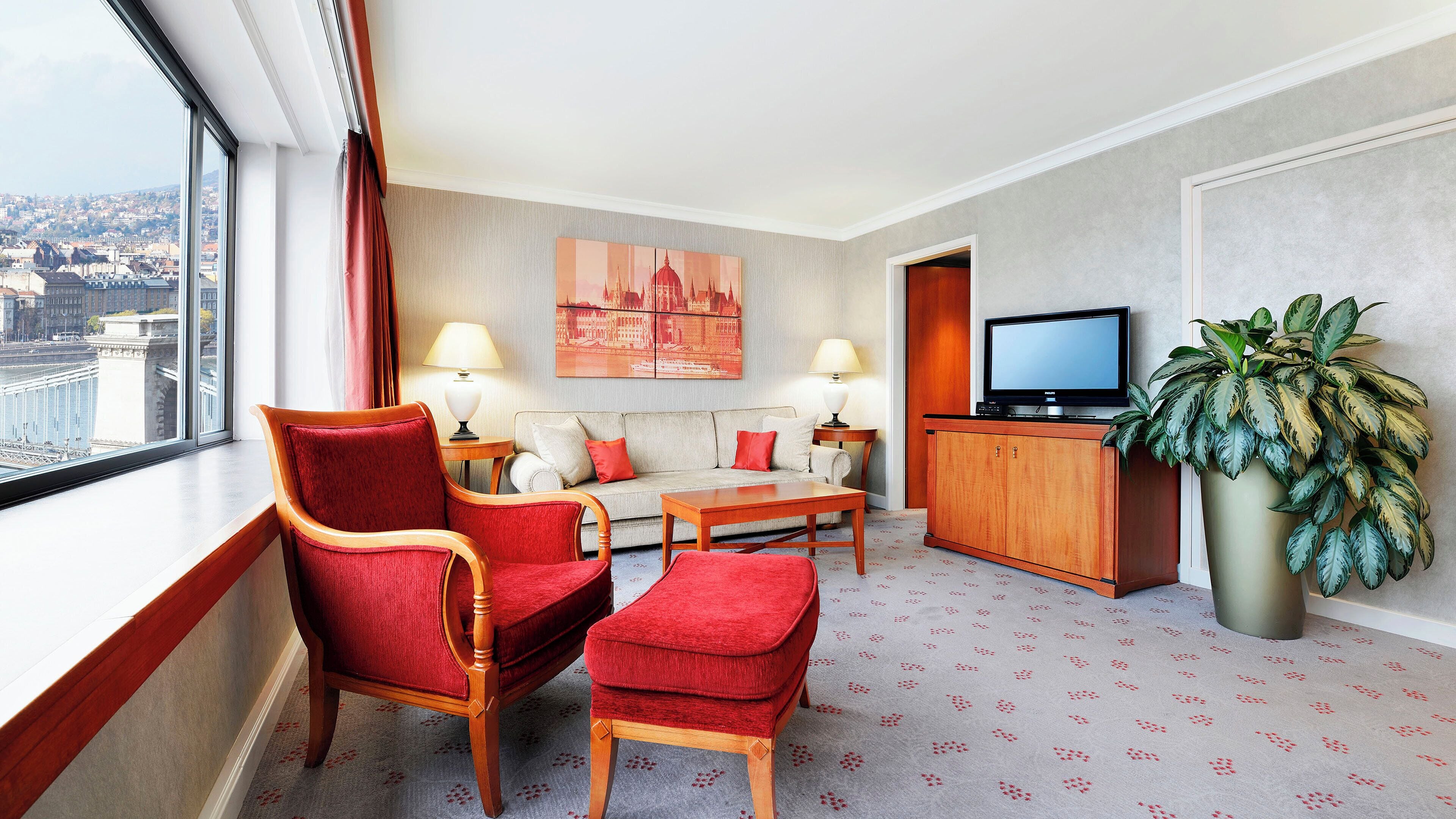 Ein Hotelzimmer mit Sofa, Sesseln, Fernseher, Lampe, Pflanze und Blick auf die Stadt.