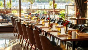 A restaurant with a long table set for dining, chairs, and a view through large windows.