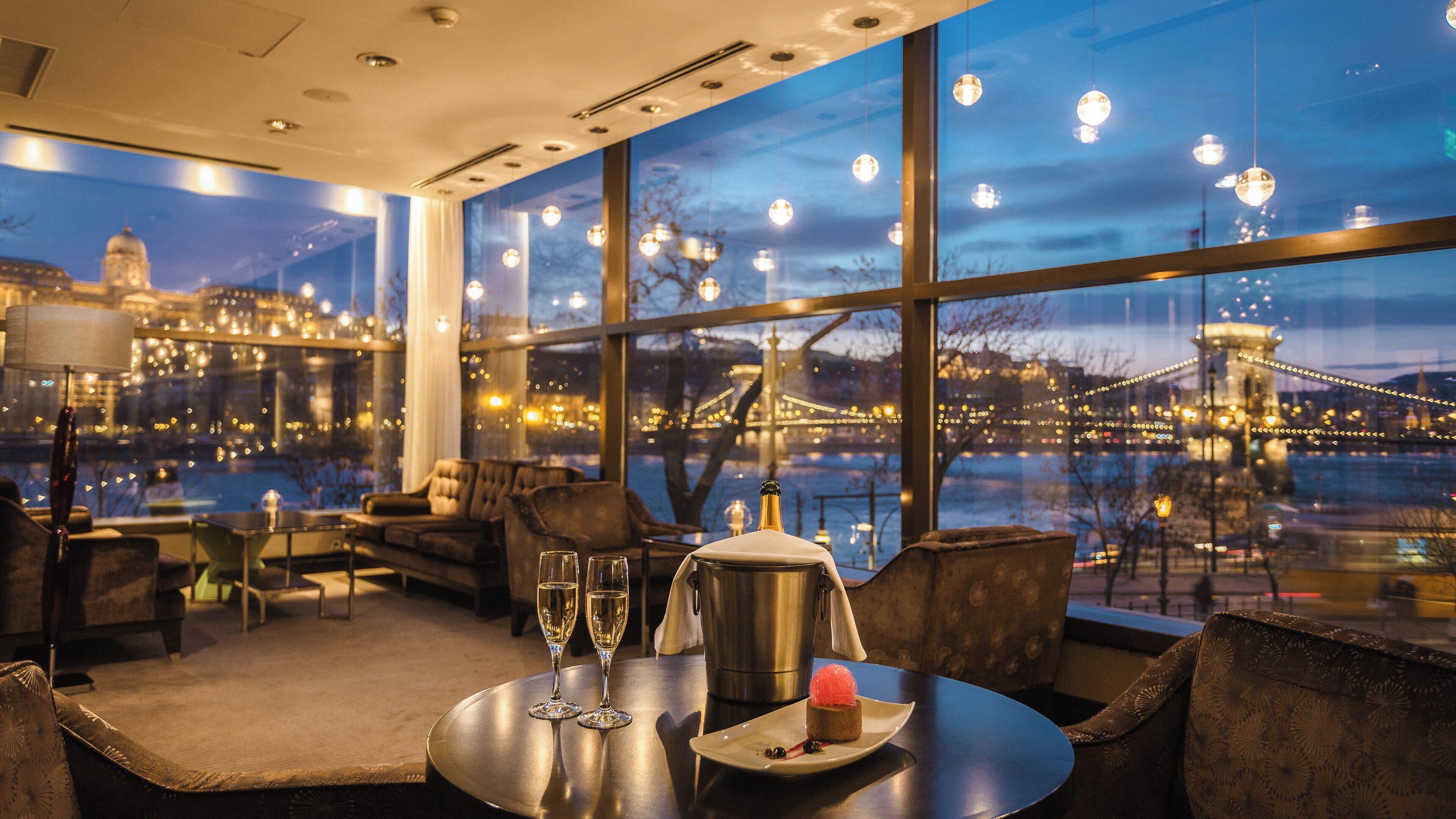Habitación de hotel moderna con un ventanal grande que ofrece vistas a la ciudad y un puente al anochecer.