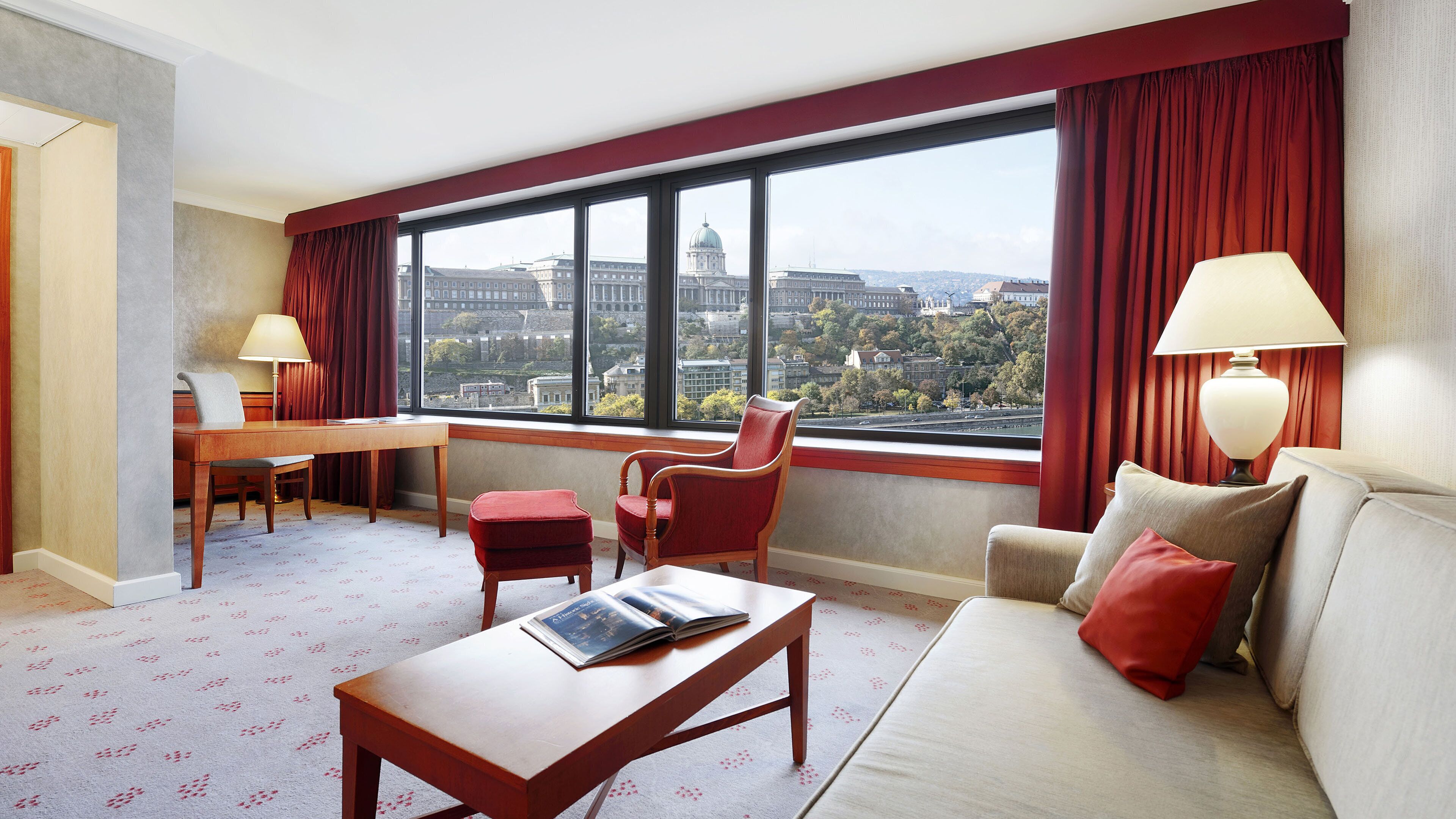 A hotel room with a large window, a desk, a chair, a sofa, and a lamp.