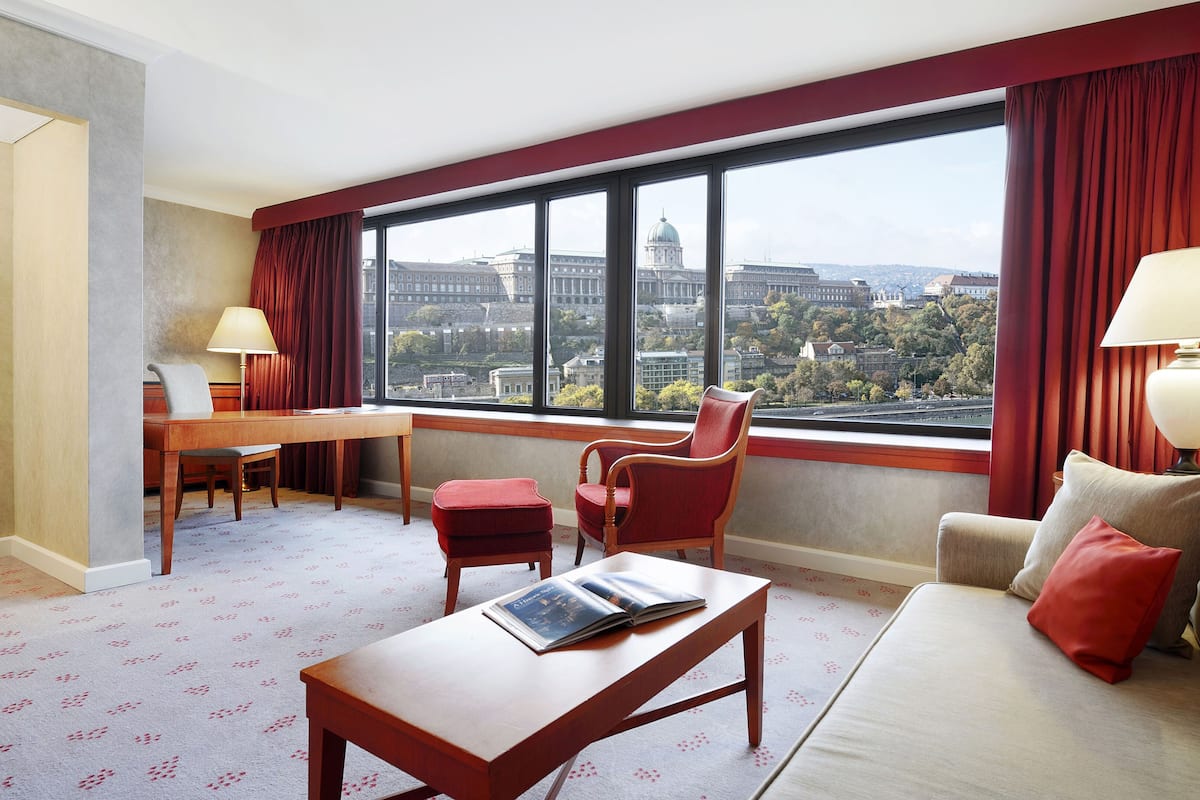 A hotel room with a large window, a desk, a chair, a sofa, and a lamp.