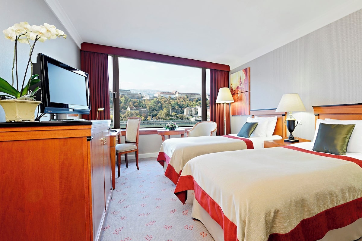 A hotel room with two beds, a desk, a chair, a TV, and a view of a cityscape.
