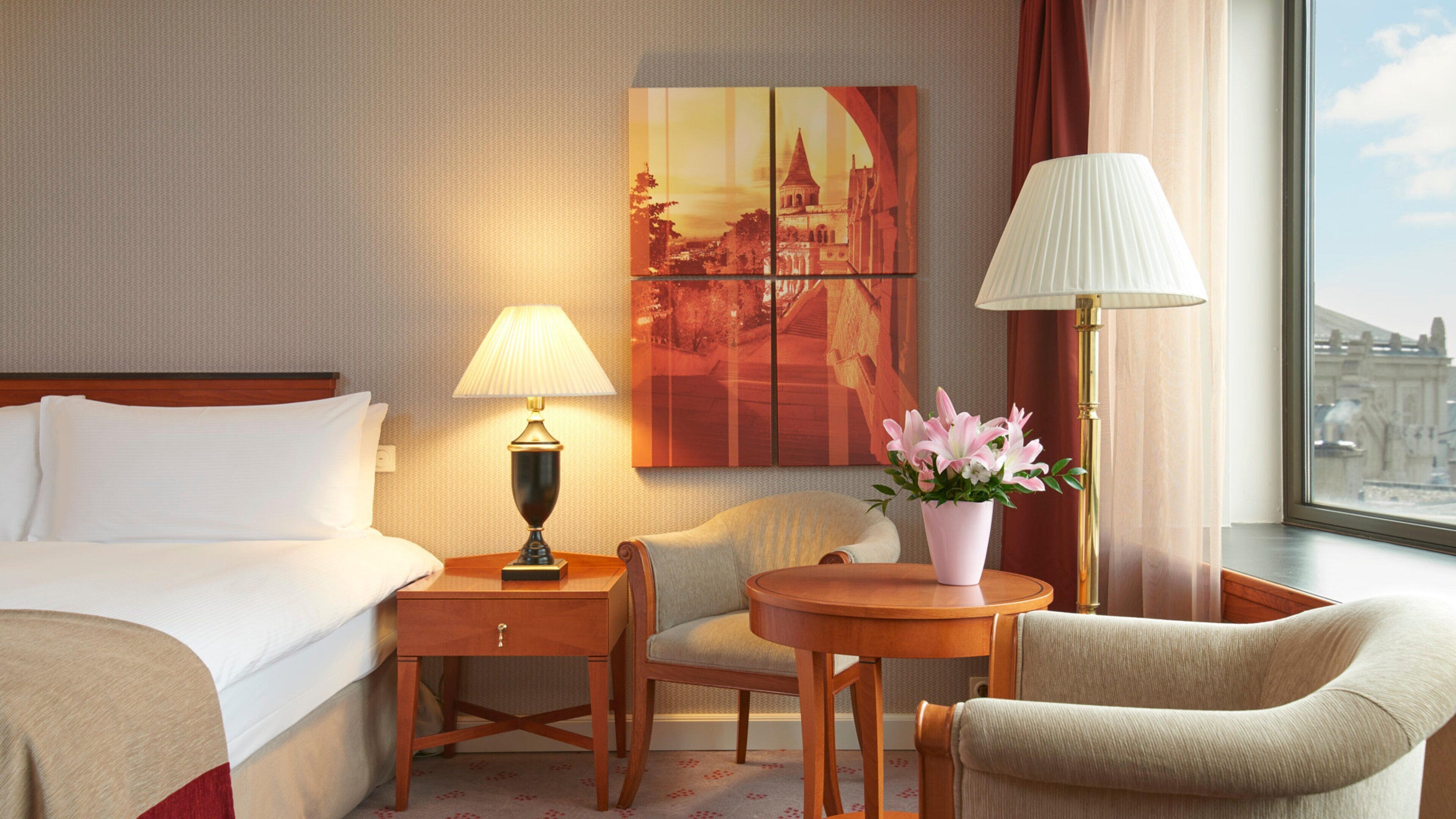 A hotel room with a bed, two chairs, a small table, a lamp, a vase with flowers, and a painting on the wall.