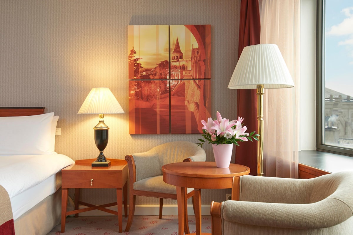 A hotel room with a bed, two chairs, a small table, a lamp, a vase with flowers, and a painting on the wall.