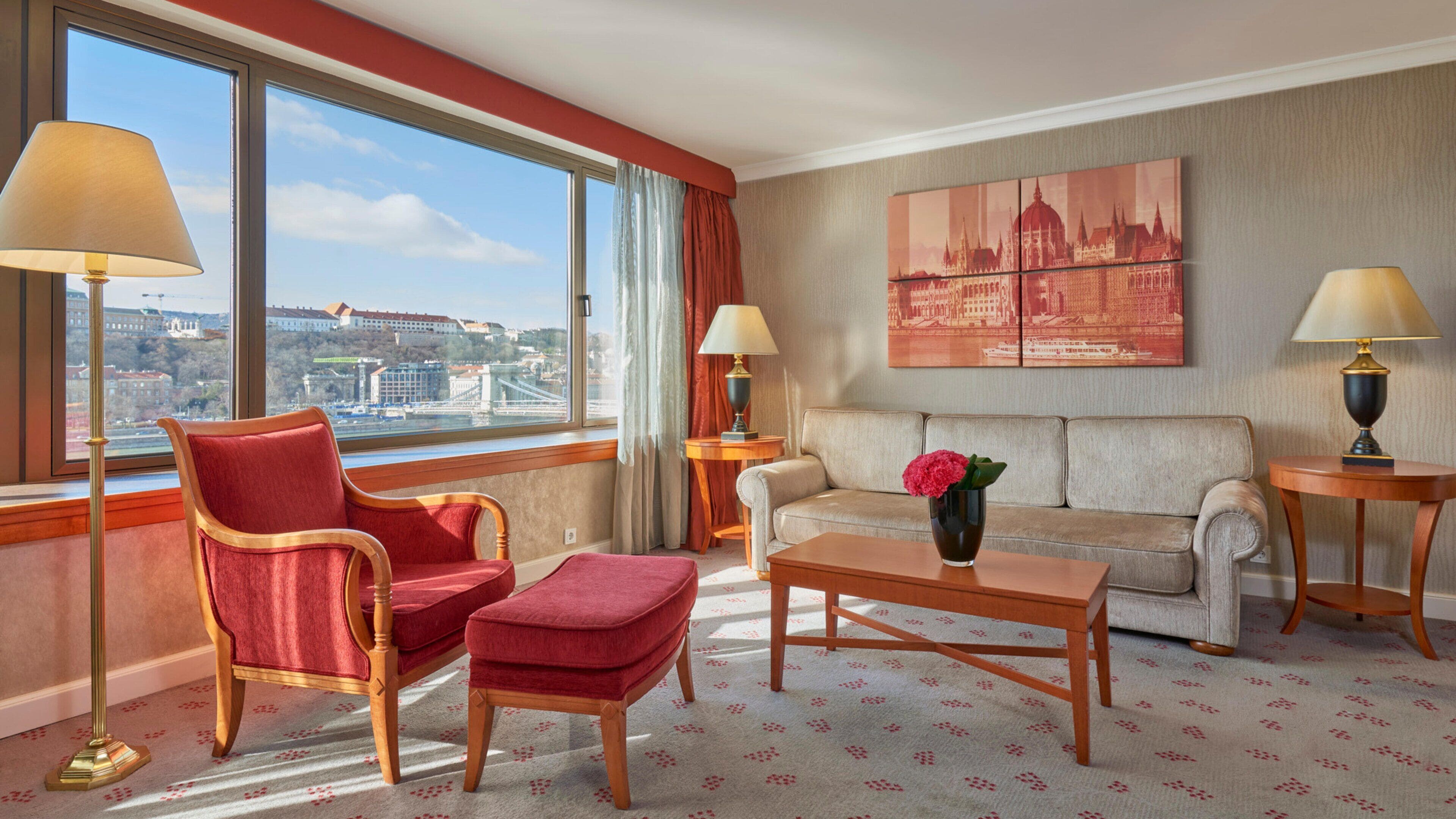 A hotel room with a large window offering a city view, a red armchair, a white sofa, and a small table with a vase of flowers.