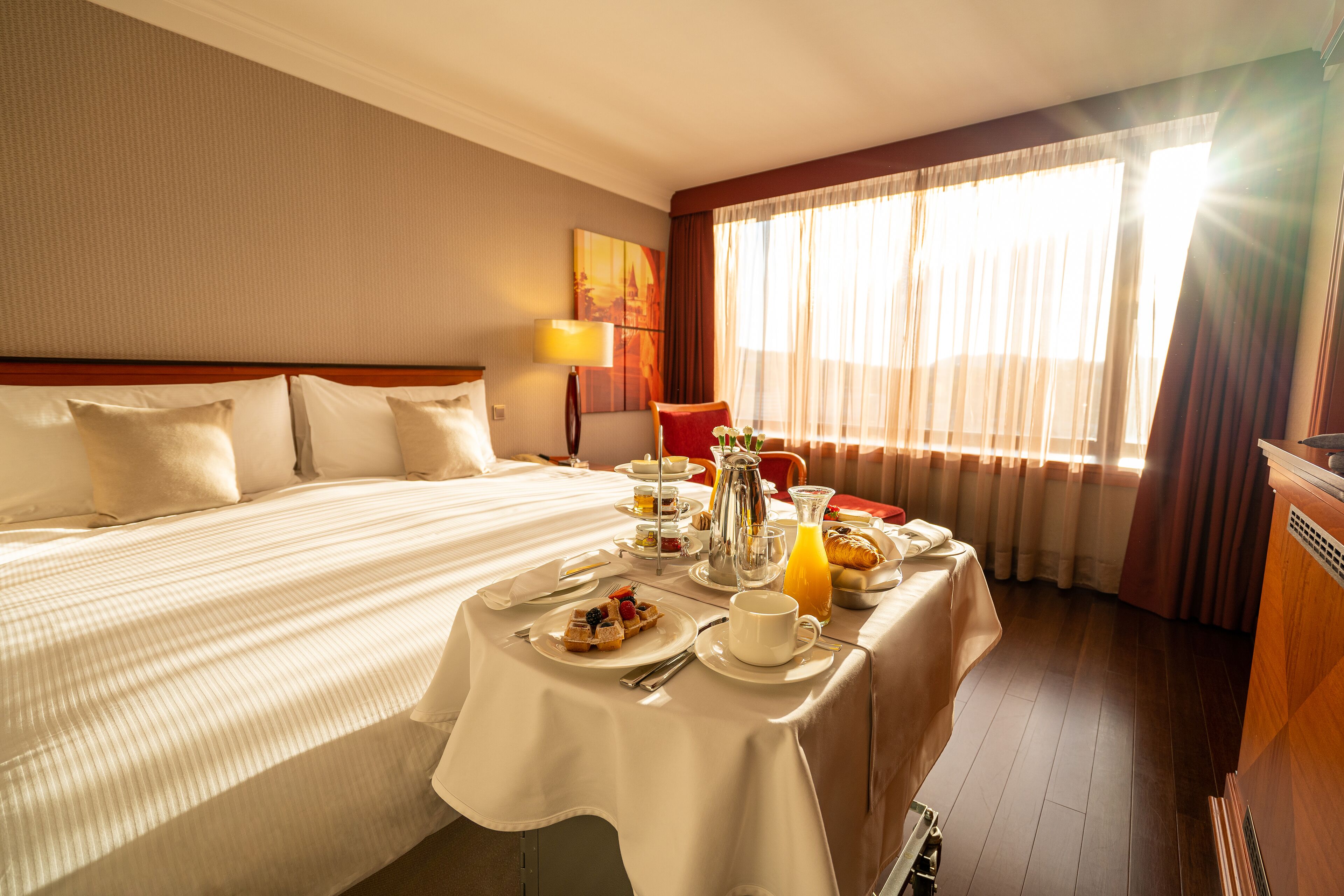 A hotel room with a neatly made bed, a breakfast table set with plates, cups, and a fruit bowl.