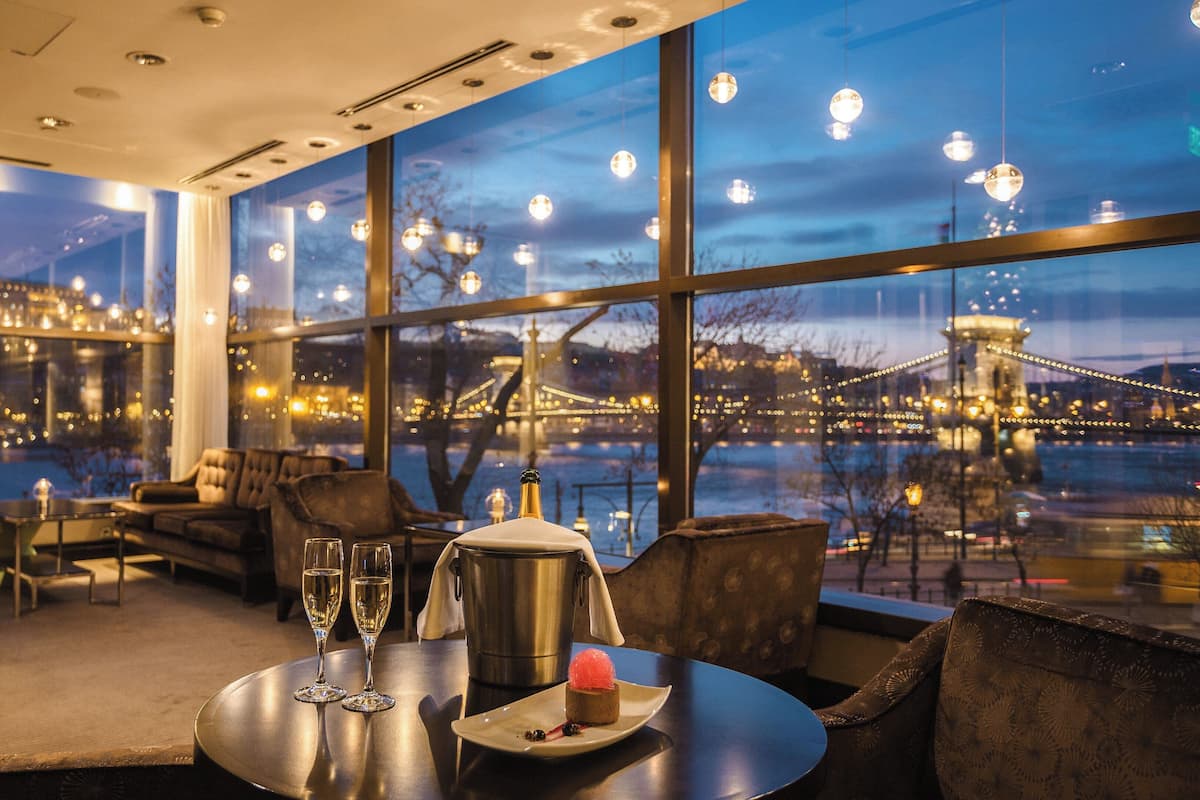 A modern hotel room with a large window overlooking a cityscape and a bridge. There is a dining area with a table set for two, a bottle of champagne, and a dessert.