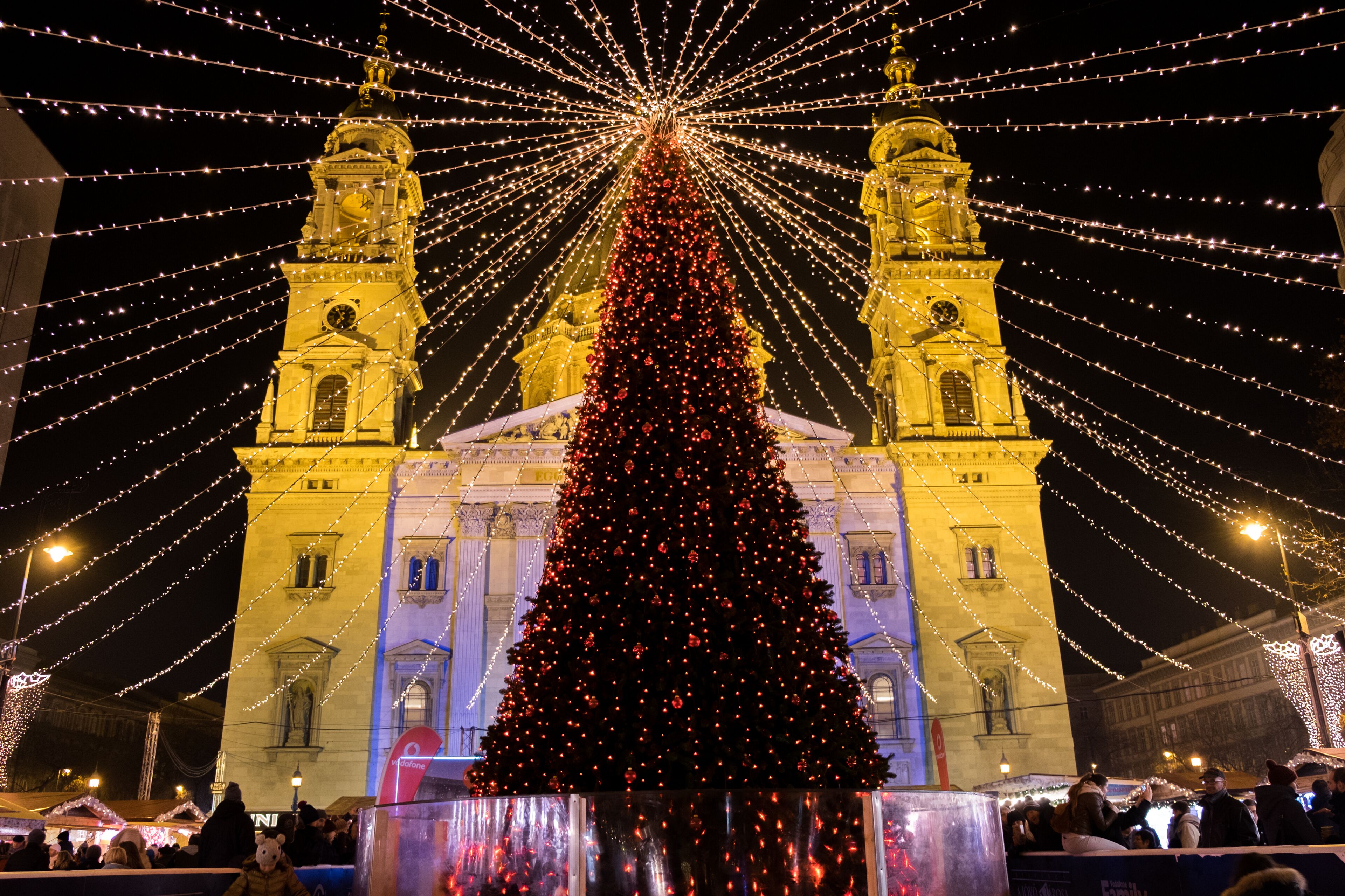 A large Christmas tree in front of a historic building with two towers, surrounded by string lights.