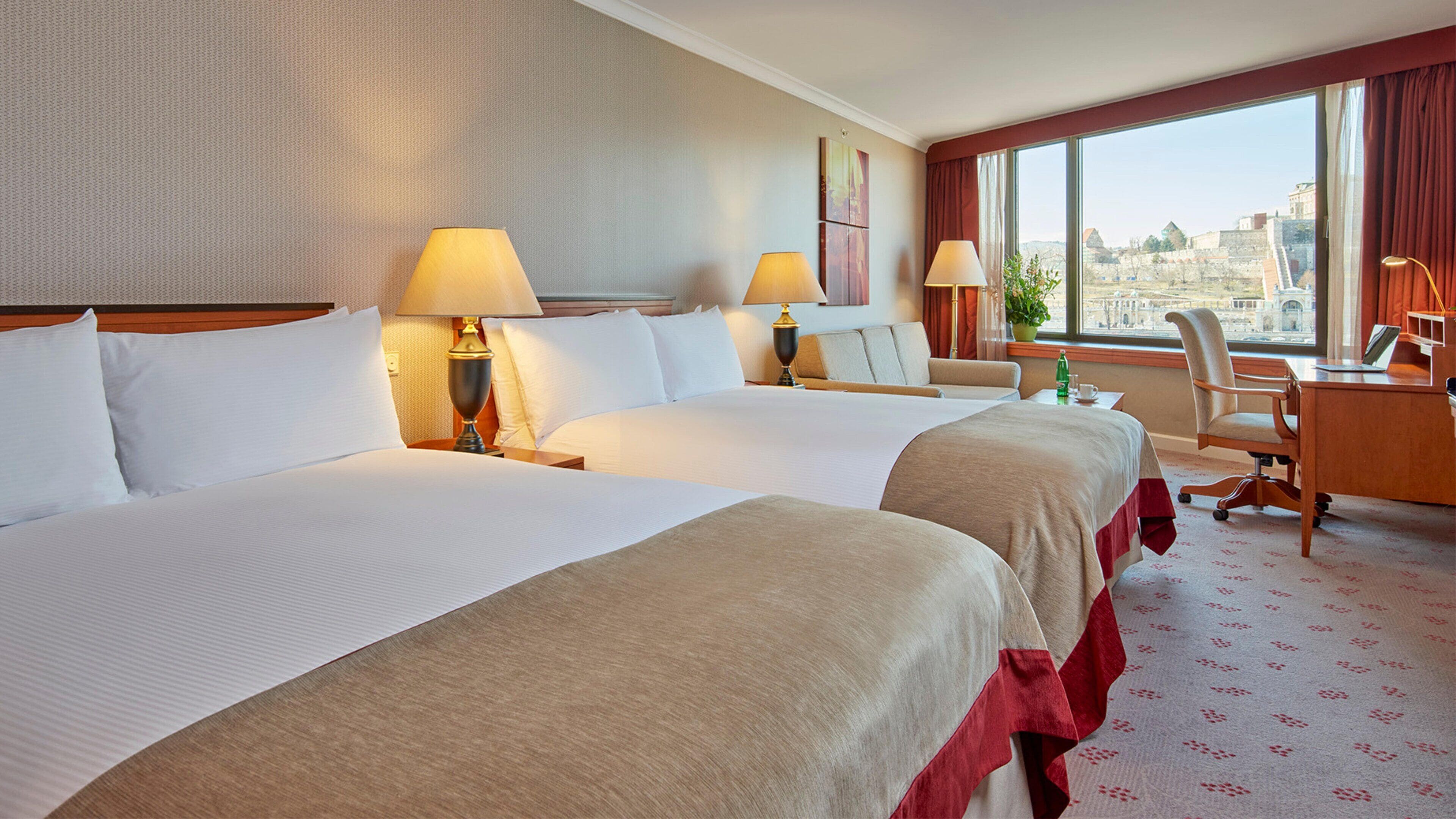 A hotel room with two beds, a desk, a chair, and a large window offering a city view.