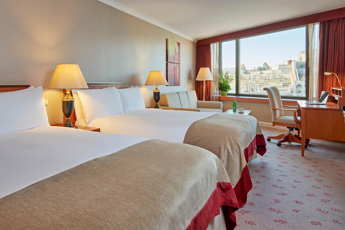 A hotel room with two beds, a desk, a chair, and a large window offering a city view.