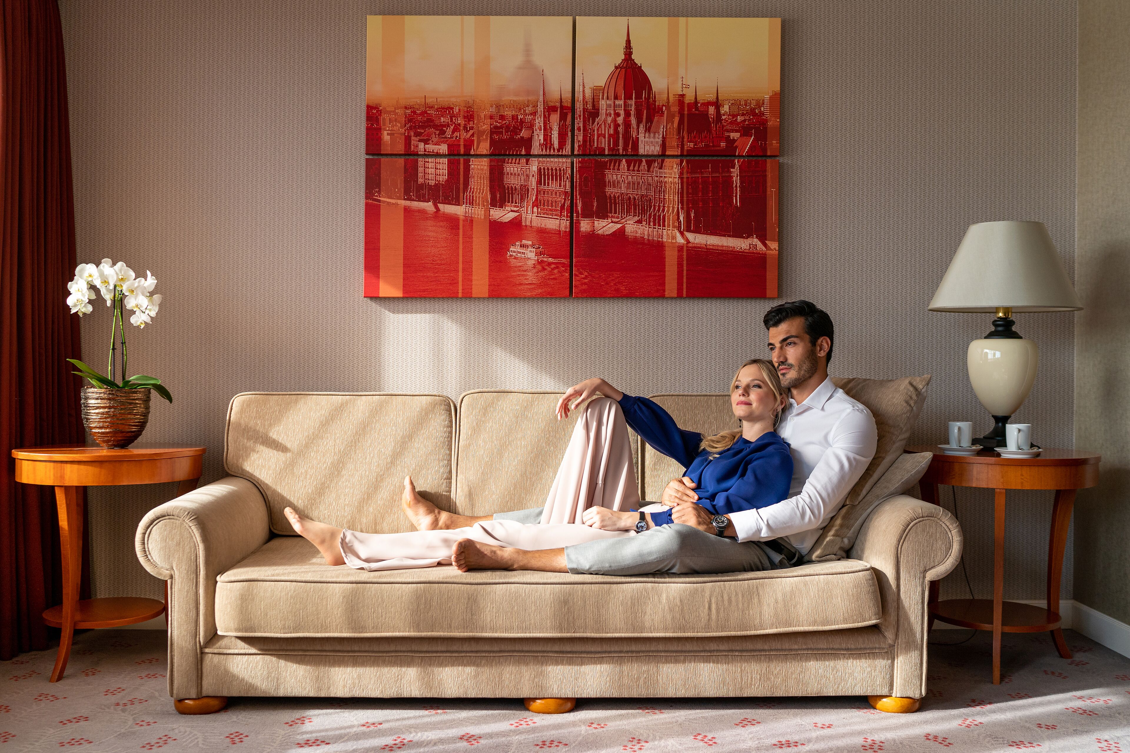 A couple on a beige sofa in a room with a cityscape painting, a lamp, and a side table with a plant.