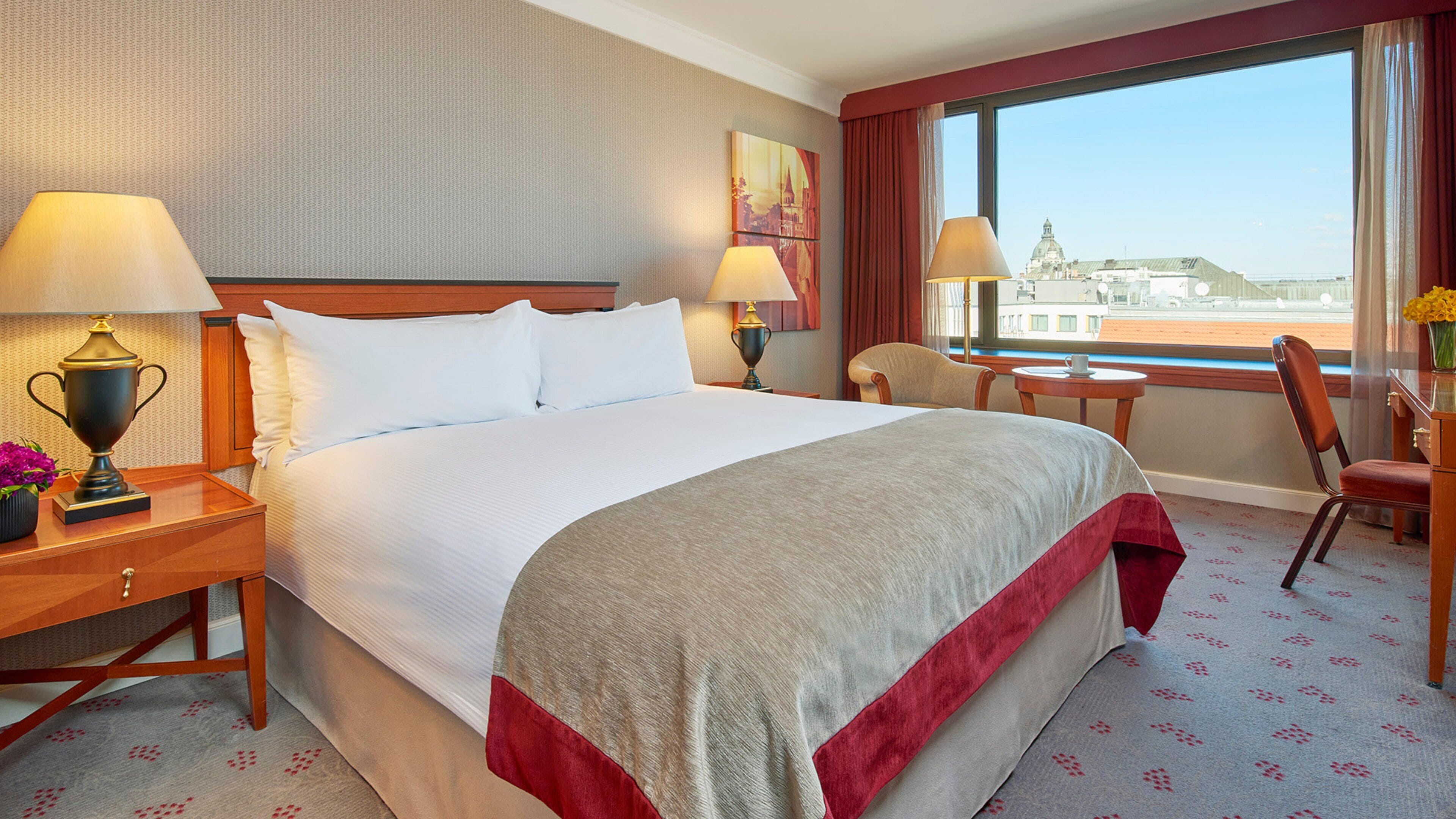 A hotel room with a large bed, two bedside tables with lamps, a chair, a small table, and a window with a view of a cityscape.