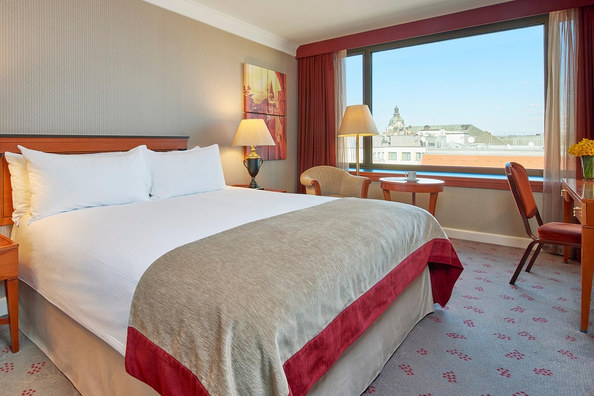 A hotel room with a large bed, two bedside tables with lamps, a chair, a small table, and a window with a view of a cityscape.