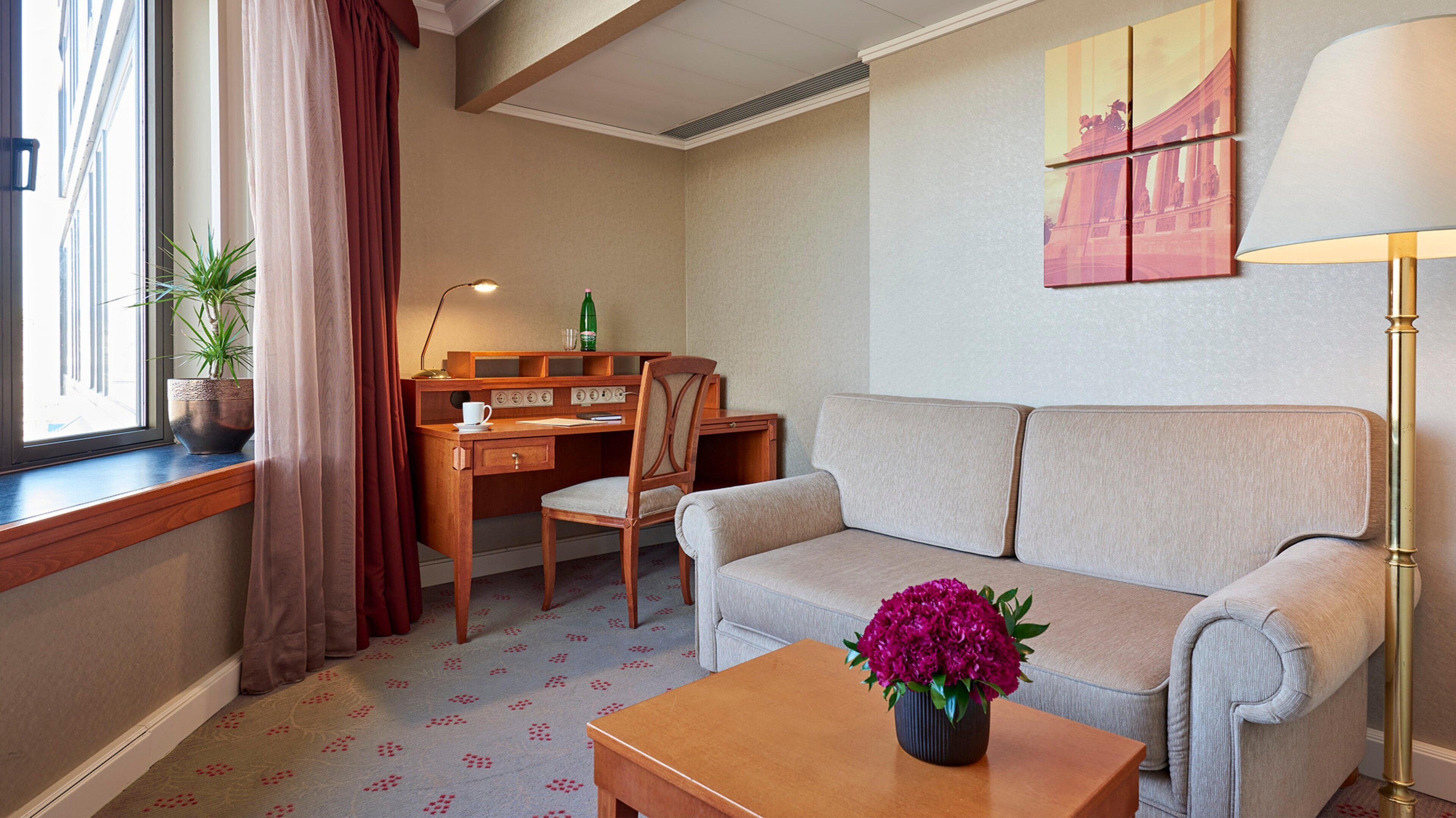 Habitación de hotel con sofá, escritorio, silla, mesa de centro, lámpara, planta en maceta y ventana con cortinas.