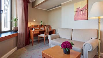 Habitación de hotel con sofá, escritorio, silla, mesa de centro, lámpara, planta en maceta y ventana con cortinas.