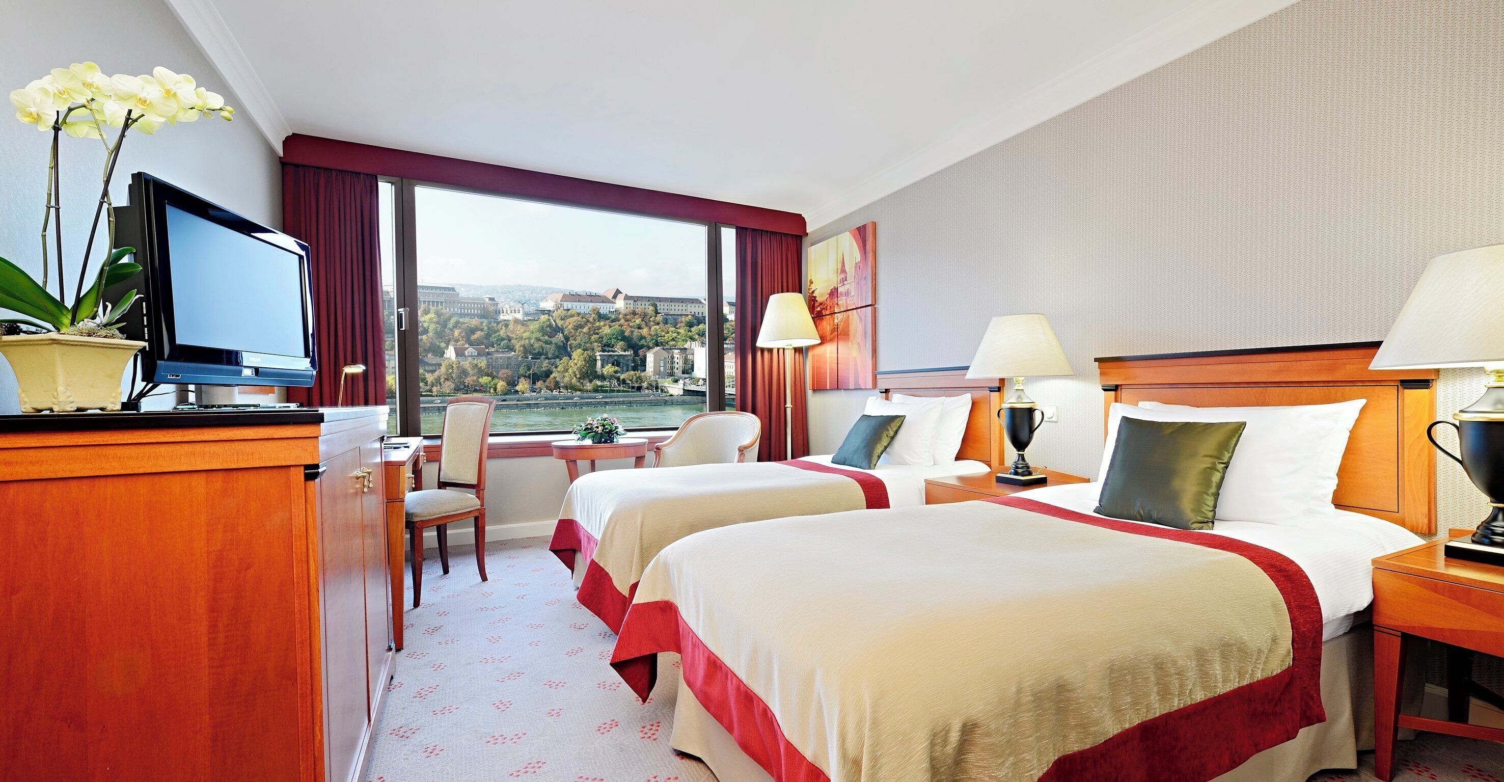 Quarto de hotel com duas camas, uma escrivaninha, uma cadeira, televisão e vista para a paisagem urbana.