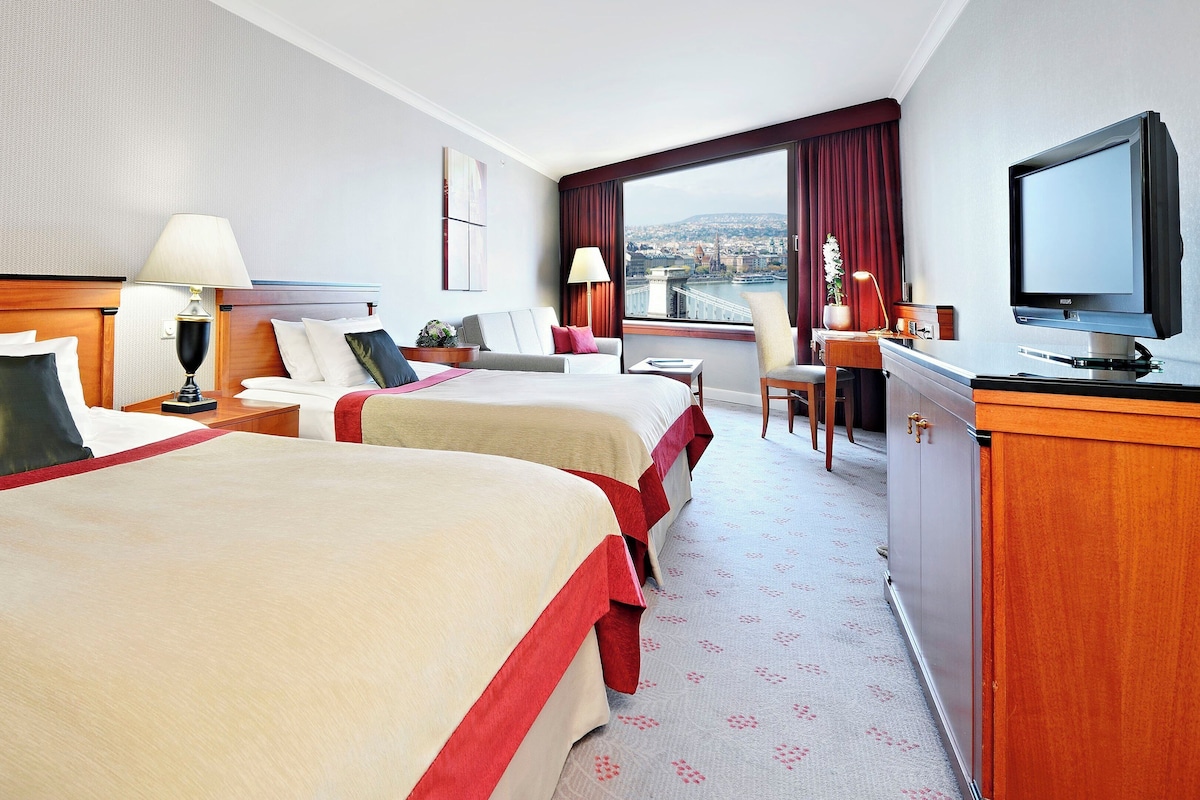 Ein Hotelzimmer mit zwei Betten, einem Schreibtisch, einem Fernseher und einem Fenster mit Blick auf die Stadt.