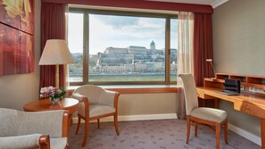 A hotel room with a large window offering a view of a historic building, a desk with a laptop, two armchairs, and a small table with a vase of flowers.
