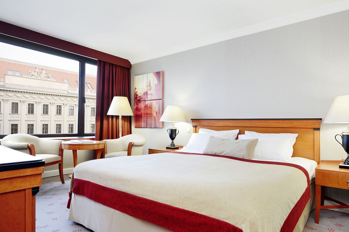 Ein Hotelzimmer mit einem großen Bett, zwei Nachttischen inklusive Lampen, einem Sessel, einem Schreibtisch und einem Fenster mit Vorhängen.