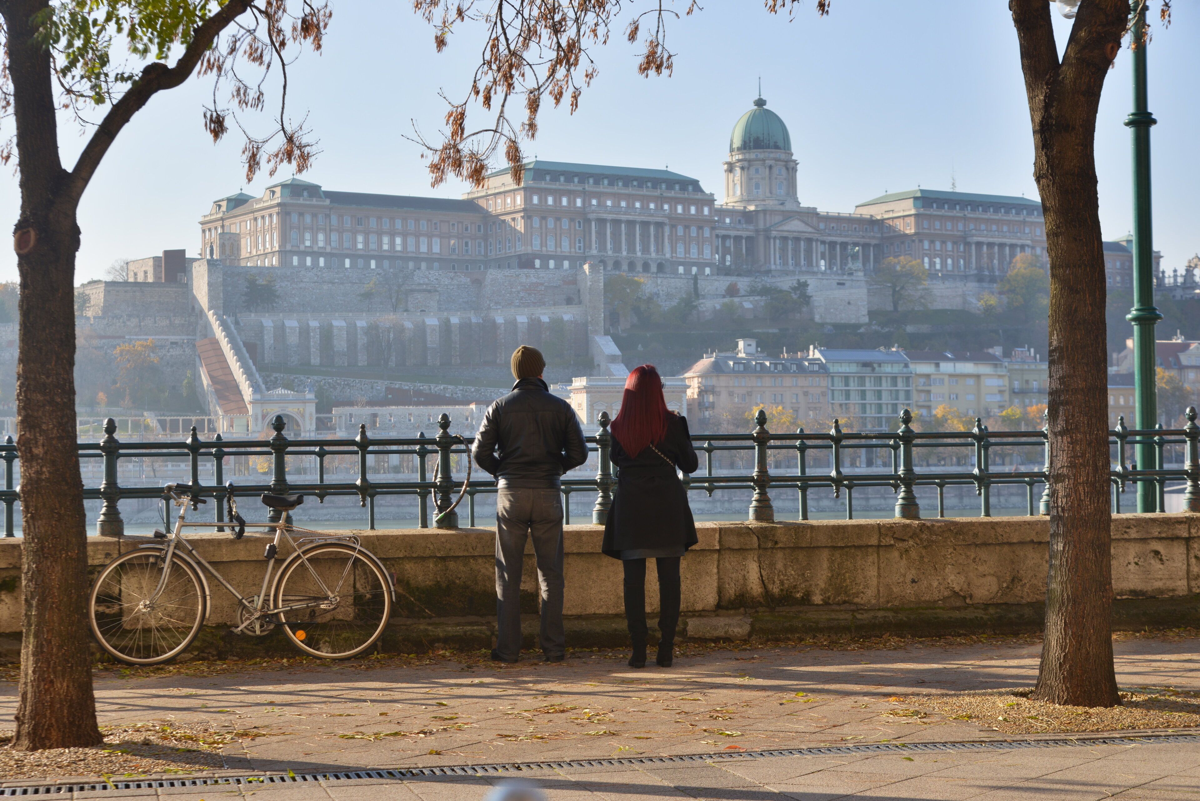 A couple standing by a river with a historic building in the background.