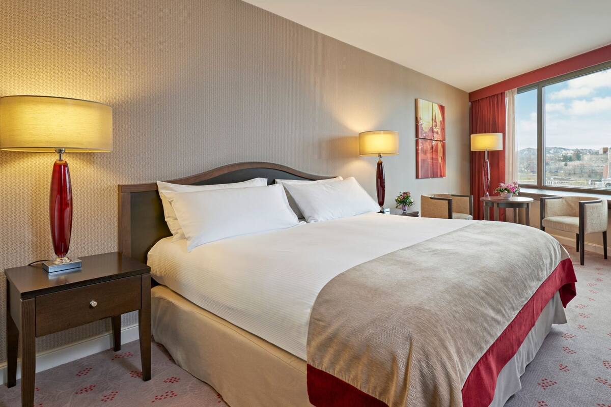 A hotel room with a large bed, two bedside tables with lamps, a desk with a chair, and a large window offering a city view.