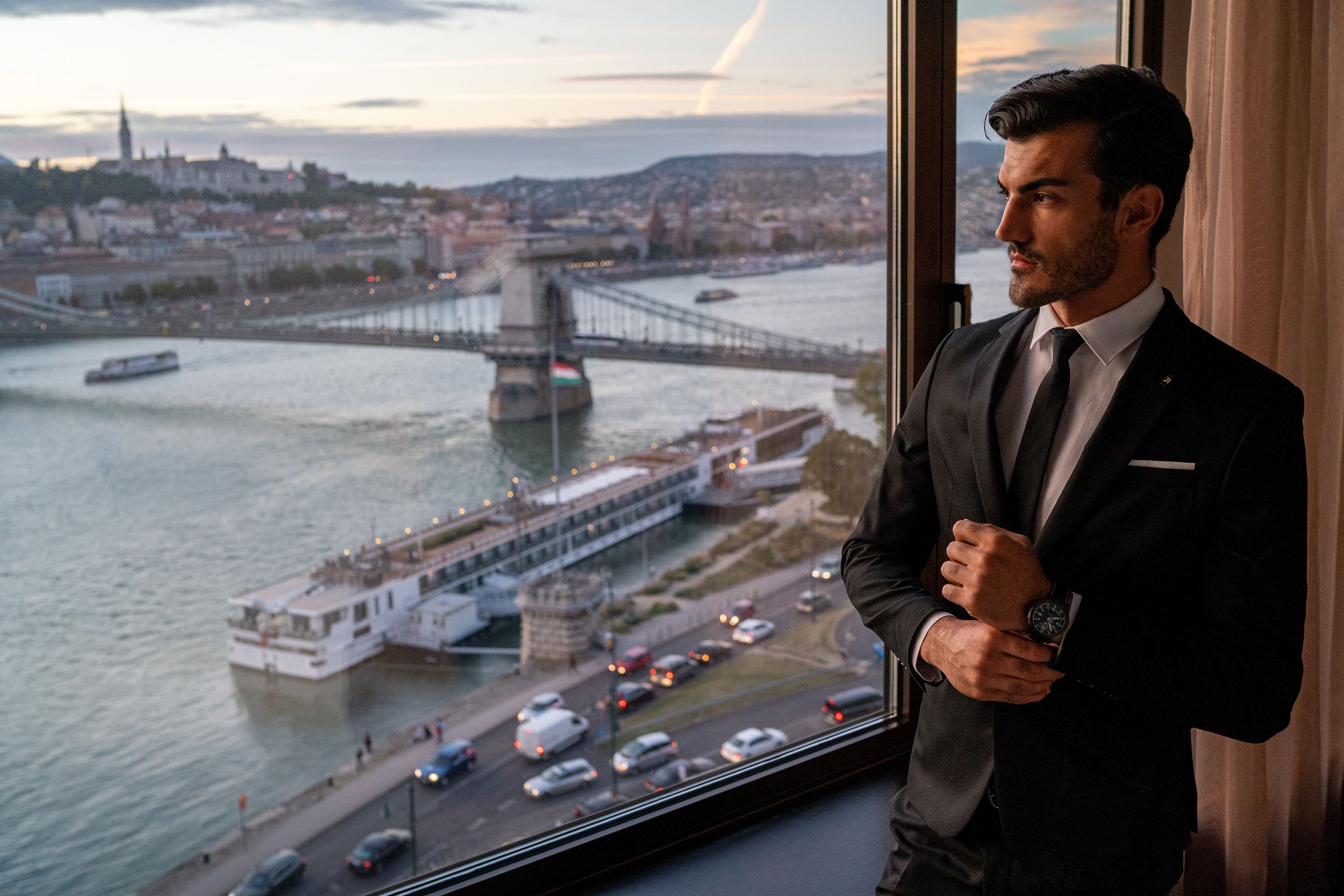 A man in a suit standing by a window overlooking a river with a bridge and a boat.