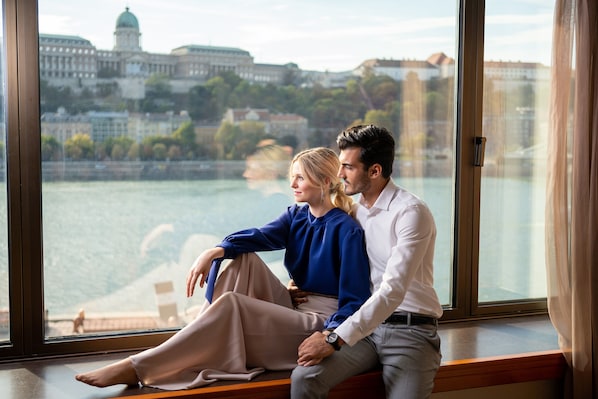 A couple sitting by a window overlooking a river with buildings in the background.
