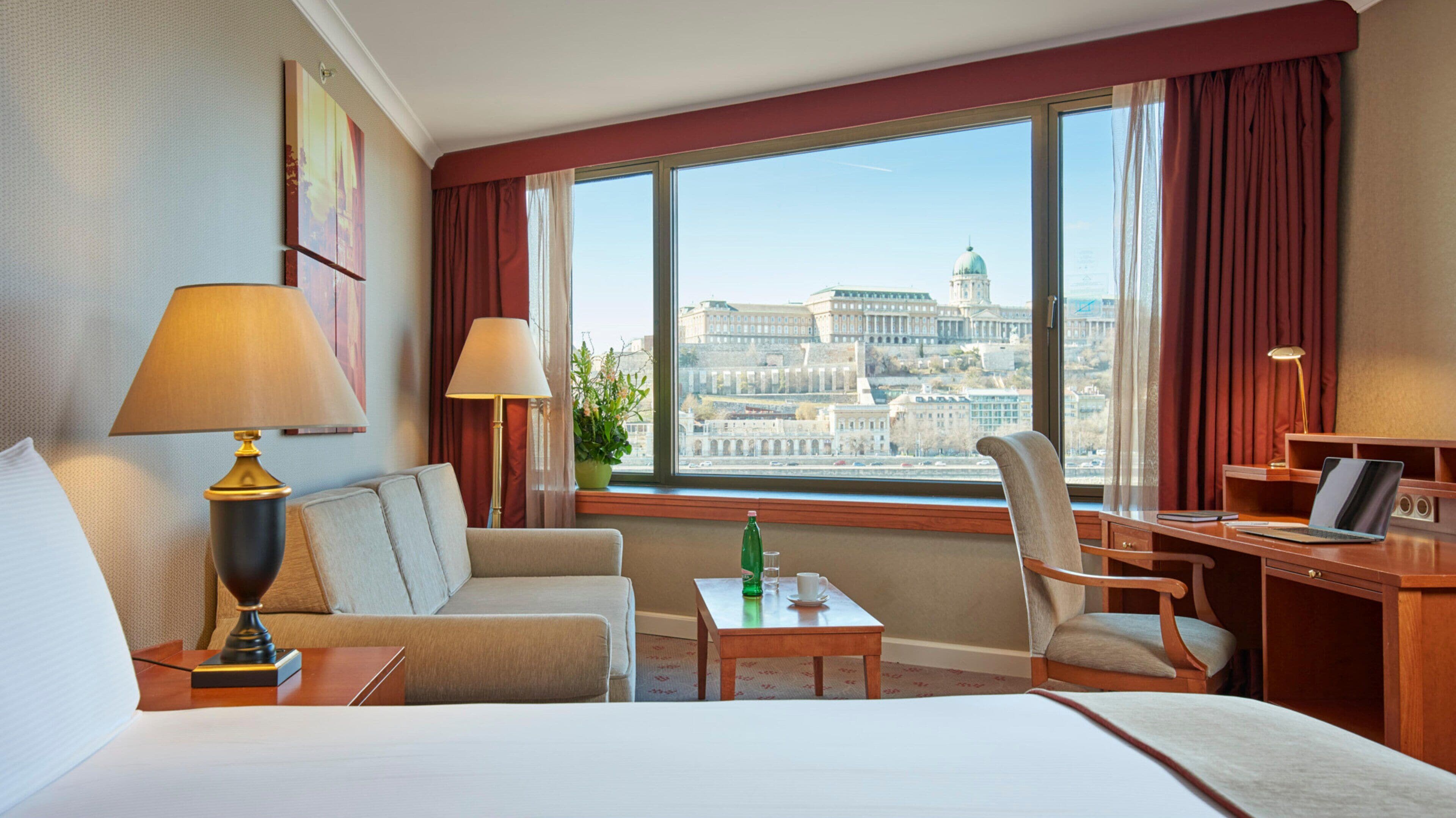Une chambre d’hôtel avec un grand lit, deux fauteuils, un bureau avec un ordinateur portable et une vue sur le paysage urbain à travers de grandes fenêtres.