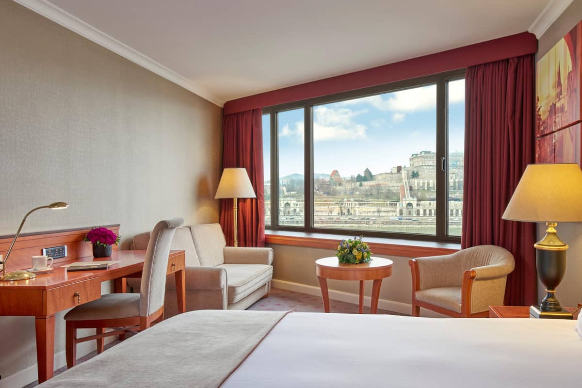 A hotel room with a bed, two armchairs, a desk, a lamp, and a view of a historic building through large windows.