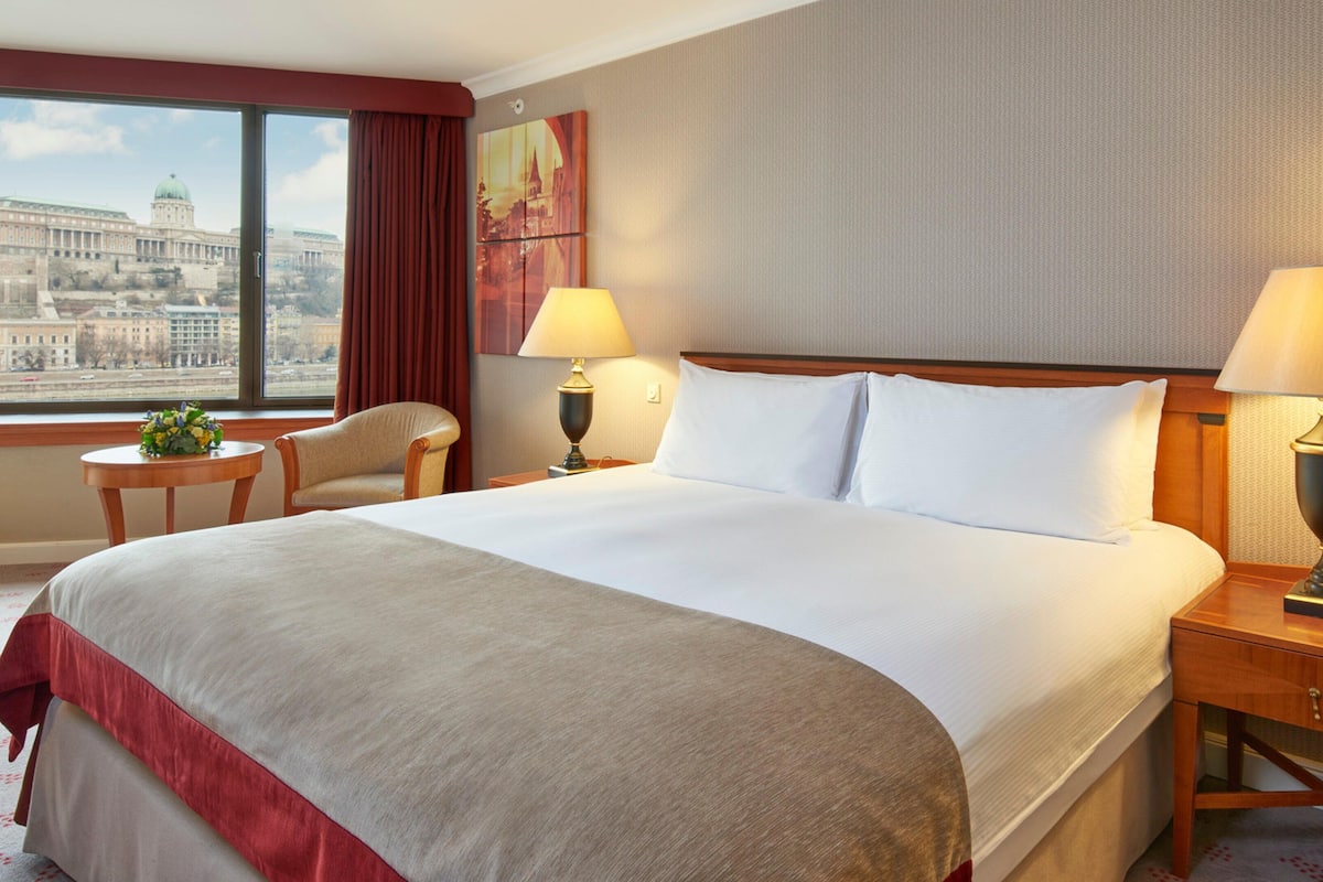 A hotel room with a large bed, two bedside tables with lamps, a chair, a window with a view of a cityscape, and a painting on the wall.