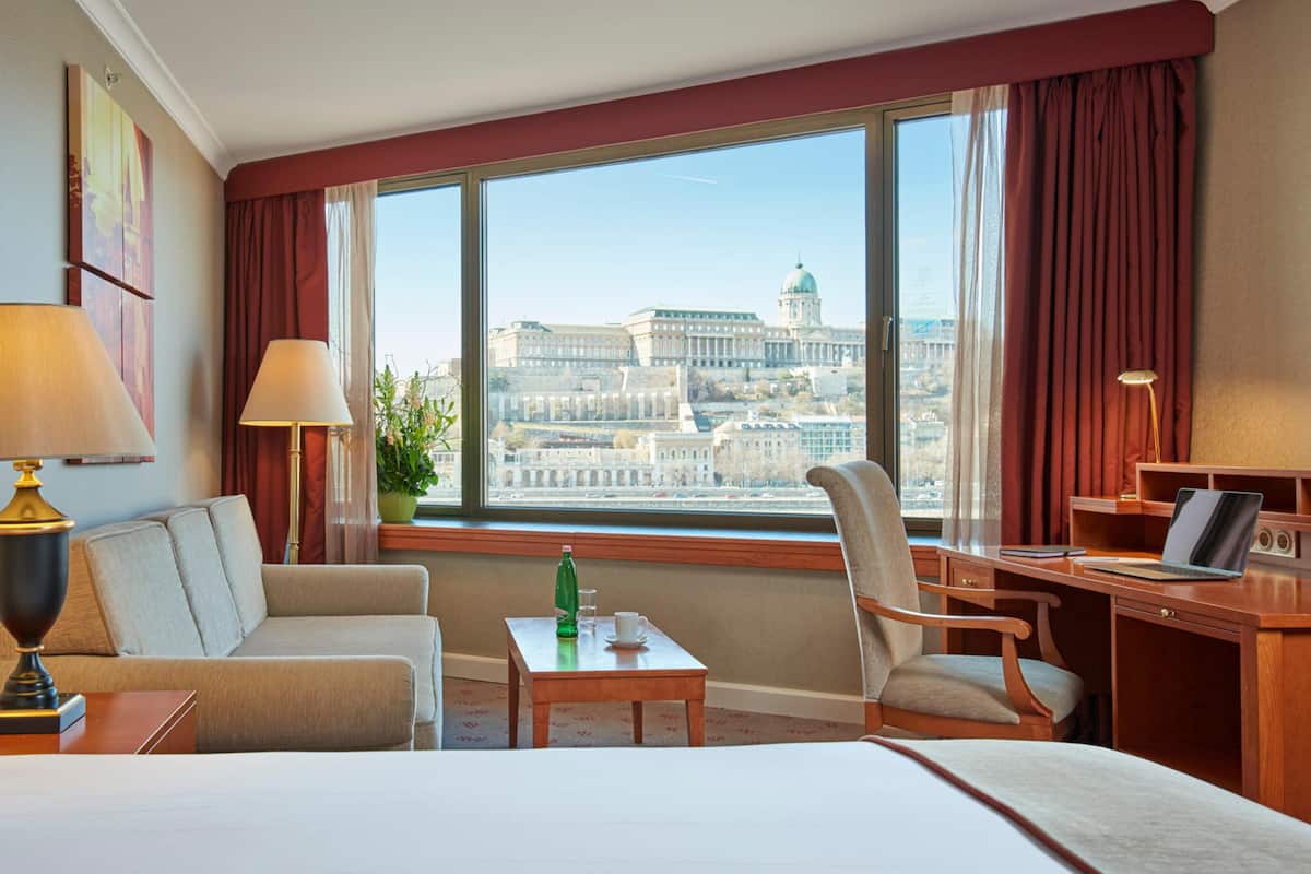 A hotel room with a large bed, two armchairs, a desk with a laptop, and a view of a cityscape through large windows.