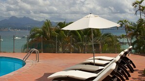 A poolside area with lounge chairs and an umbrella, overlooking a body of water and mountains. - Hotel Bakoua Les Trois Ilets (Trois-Ilets)